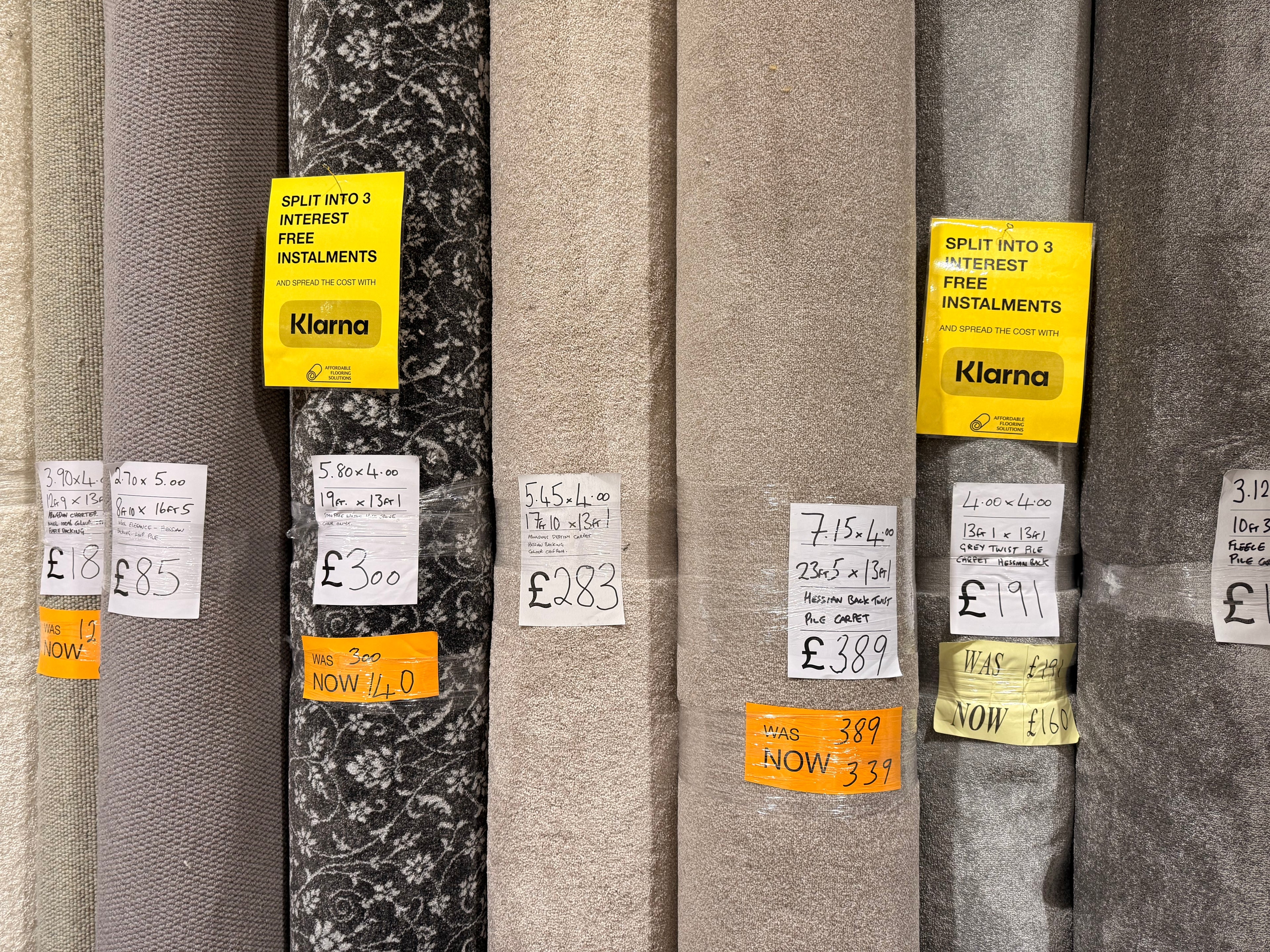 A row of carpet rolls and remnants on display at Affordable Flooring Solutions in Bury St Edmunds, each labelled with sizes and prices, showing various grey and beige styles with sale and Klarna signage visible.